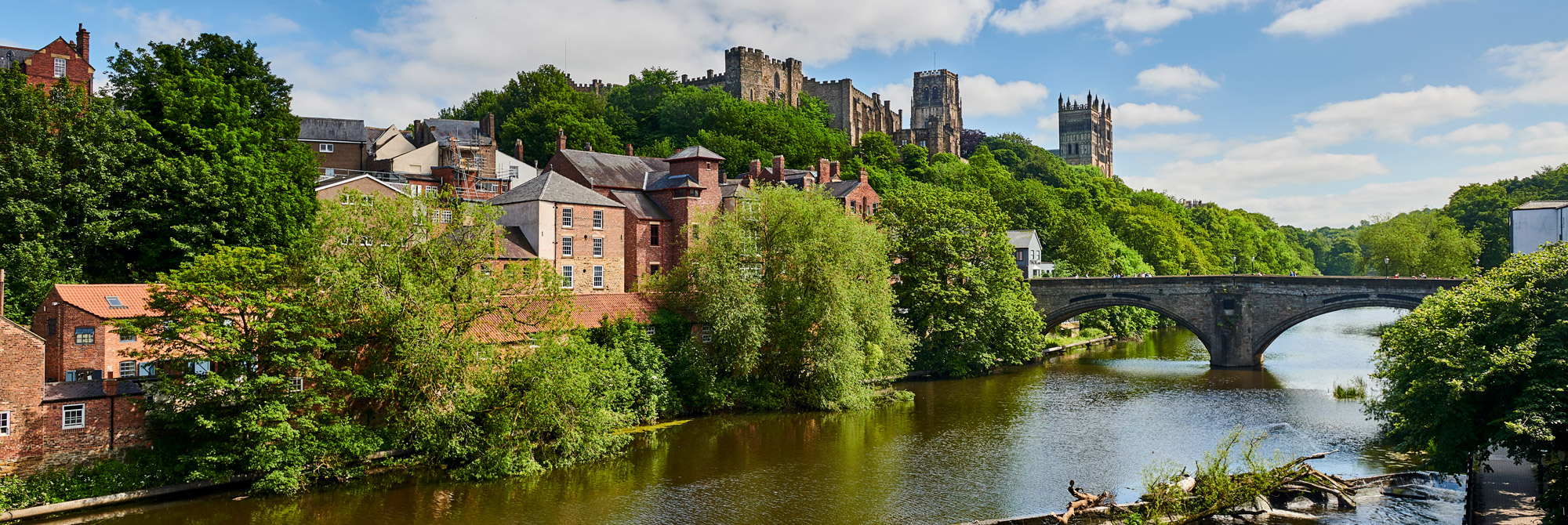 Photograph of Durham Cathedral and Castle high above the river, with green trees on the riverbanks and Framwellgate Bridge in the foreground